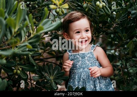 Happy little girl standing in public park against green leaves
