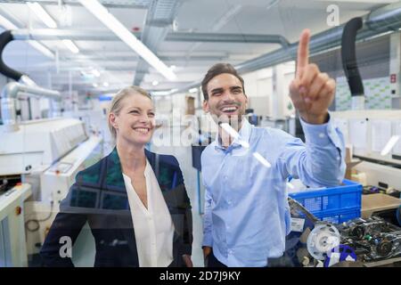 Happy young male technician pointing at graphical interface on glass in factory Stock Photo