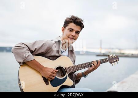 Handsome young man playing guitar while practicing at promenade against sky Stock Photo