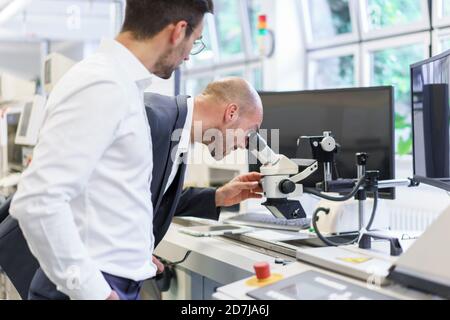 Young male technician standing by businessman looking through microscope at laboratory Stock Photo
