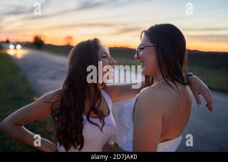 Smiling female friends looking at each other while standing on road against sky at sunset Stock Photo