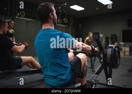 Man using rowing machine with people exercising in background at gym Stock Photo