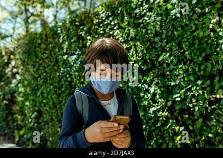 Schoolboy wearing mask using mobile phone while standing by plants Stock Photo