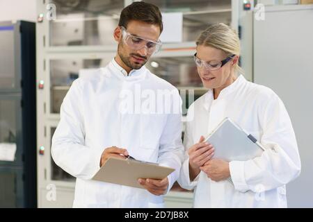 Young male technician discussing over clipboard with female blond colleague holding digital tablet at laboratory Stock Photo