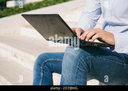 Man typing on laptop while sitting outdoors during sunny day Stock Photo