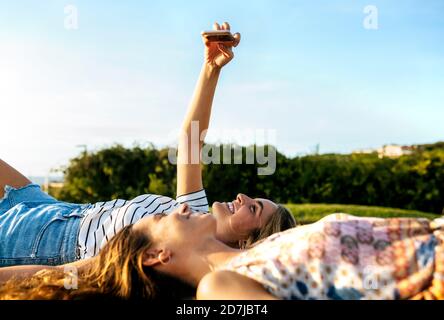 Smiling young woman taking selfie with female friend while relaxing on field during sunny day Stock Photo