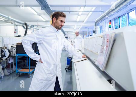 Young male technician in lab coat looking at reports on machinery Stock Photo