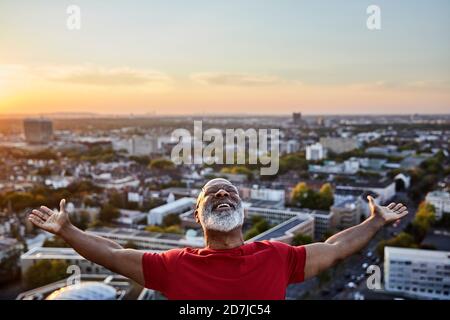 Happy bearded man with arms outstretched while standing on building terrace in city during sunset Stock Photo