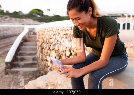 Smiling beautiful Hispanic woman using smart phone while sitting outdoors Stock Photo