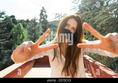 Young woman wearing protective face mask doing hand gesture while standing in balcony Stock Photo