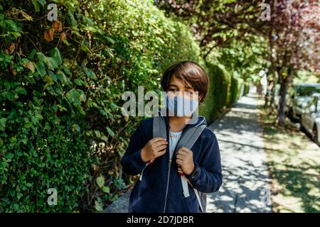 Schoolboy wearing mask looking away while standing by plants on footpath Stock Photo