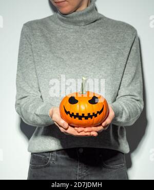 Crop unrecognizable woman holding halloween scary face bright orange pumpkin. Young woman in gray cashmere sweater and black jeans with jack-o-lantern pumpkin in hands. Snapshot style.. Copy space Stock Photo