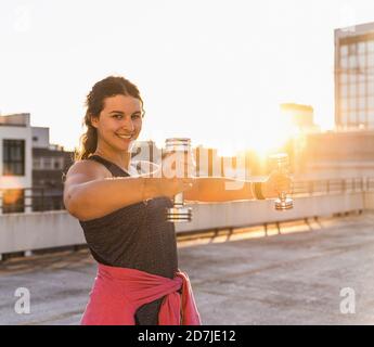 Smiling young woman exercising with dumbbells on terrace against clear sky at sunset Stock Photo