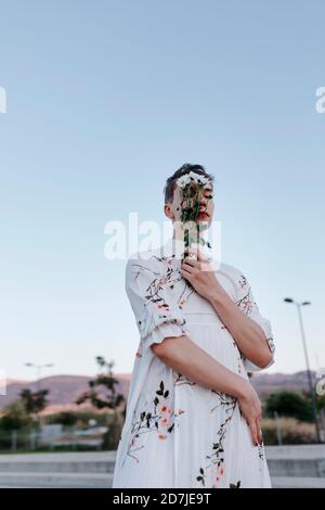 Gender fluid man covering face with bouquet of daisies while standing on street during sunset Stock Photo
