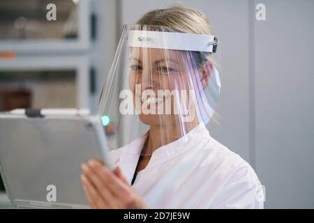 Happy mature female scientist wearing protective face shield while using digital tablet at laboratory Stock Photo