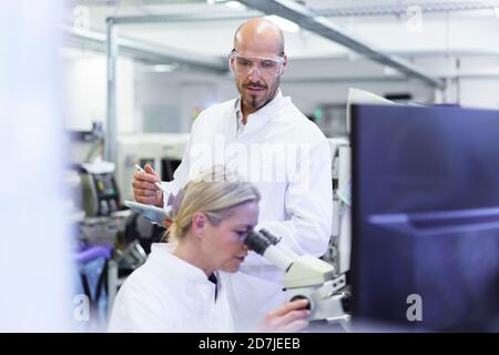 Male technician looking at female scientist doing research through microscope in laboratory Stock Photo