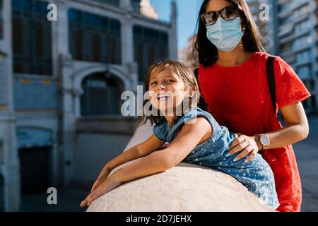 Mother wearing mask standing with daughter by retaining wall in city Stock Photo