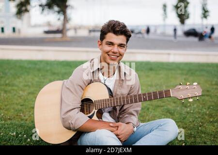 Smiling handsome young man sitting with guitar on grass at park Stock Photo
