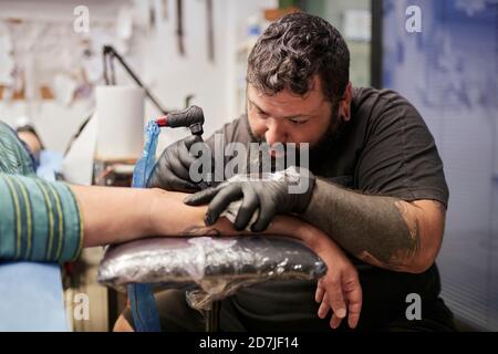 Bearded artist tattooing on male customer's hand in studio Stock Photo