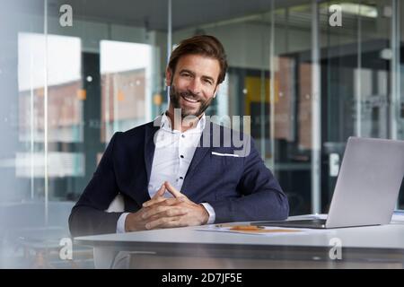 Smiling businessman with hands clasped sitting by desk in office