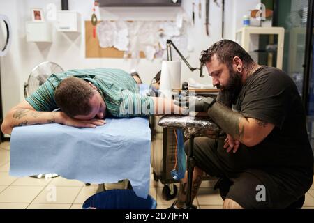 Bearded artist tattooing on male customer's hand lying on bed in studio Stock Photo