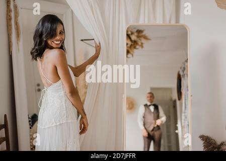 Reflection of bridegroom in mirror while bride looking over shoulder at wedding dress shop Stock Photo