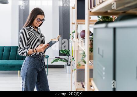 Smiling young female architect reading book while standing by rack at workplace Stock Photo
