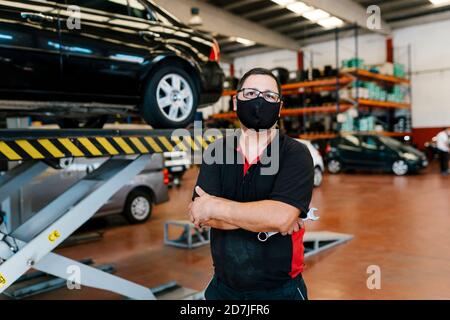Male mechanic wearing mask with arms crossed standing in workshop Stock Photo