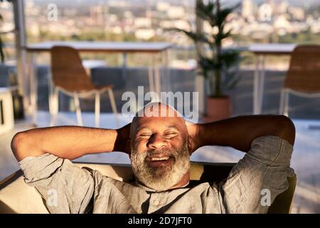 Smiling mature man relaxing on sofa at building terrace during sunset Stock Photo