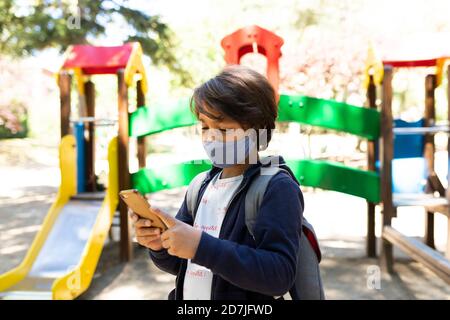 Schoolboy wearing mask using smart phone while standing in schoolyard Stock Photo