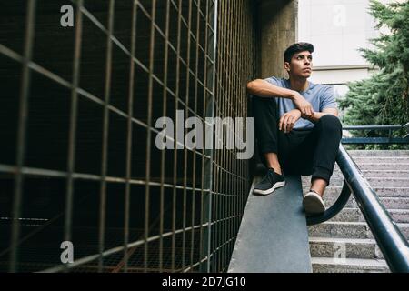 Thoughtful young man sitting on retaining wall by fence Stock Photo