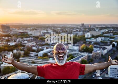 Bearded man shouting while standing with arms outstretched on building terrace in city during sunset Stock Photo