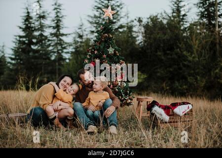 Smiling family sitting by Christmas tree on grassy land at countryside during sunset Stock Photo