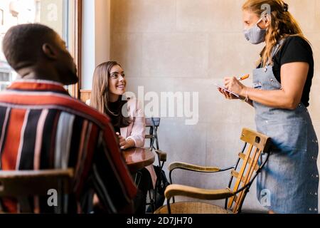 Waitress in protective face mask taking order from young couple at cafe Stock Photo