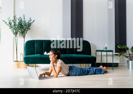 Thoughtful female architect looking away while lying with laptop on floor by sofa in creative workplace Stock Photo