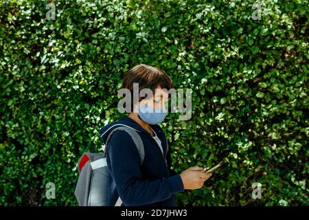 Schoolboy wearing mask using smart phone while standing against plants Stock Photo