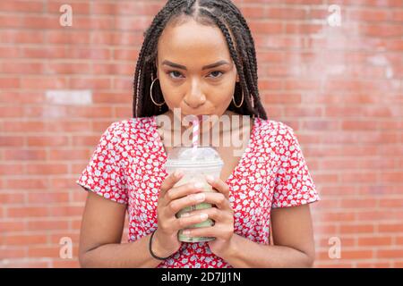 Young woman drinking smoothie while standing against wall Stock Photo