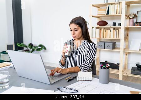Confident beautiful businesswoman using laptop while drinking at desk in office Stock Photo