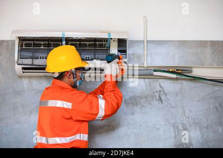 Air conditioner technician mechanic wearing mask and helmet to prevent disease, Covid 19 Currently using electric drill to install air conditioner. Stock Photo
