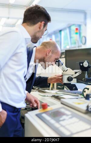Young technician standing by male colleague looking through microscope at laboratory Stock Photo