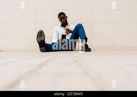 Businessman sitting with smart phone and reusable cup against wall on footpath Stock Photo