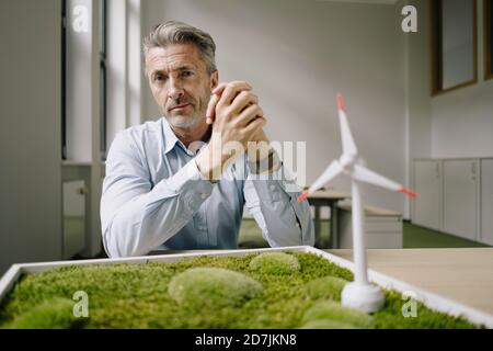 Businessman with hands clasped sitting by wind turbine toy and moss frame on table at office Stock Photo