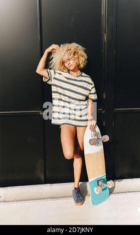 Blond woman with Afro hairstyle holding skateboard while standing against black wall Stock Photo