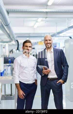 Smiling mature businessman holding digital tablet while standing by young male engineer at factory Stock Photo