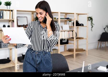 Smiling young female architect talking on smart phone while looking at plan in office Stock Photo