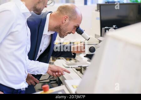Young technician pointing to male colleague looking through microscope at laboratory Stock Photo