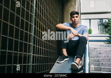 Serious young man sitting on retaining wall by fence Stock Photo