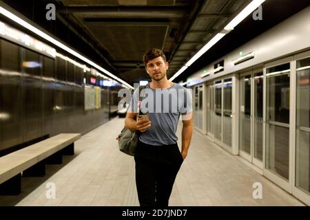 Handsome young man holding smart phone while walking with hand in pocket at illuminated subway platform Stock Photo