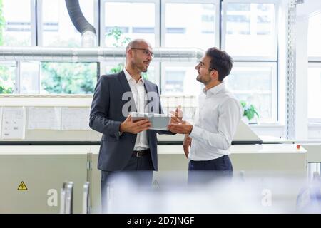 Male professionals discussing over digital tablet while standing against window at factory Stock Photo