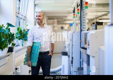Smiling male engineer looking away while holding large circuit board by machinery at factory Stock Photo